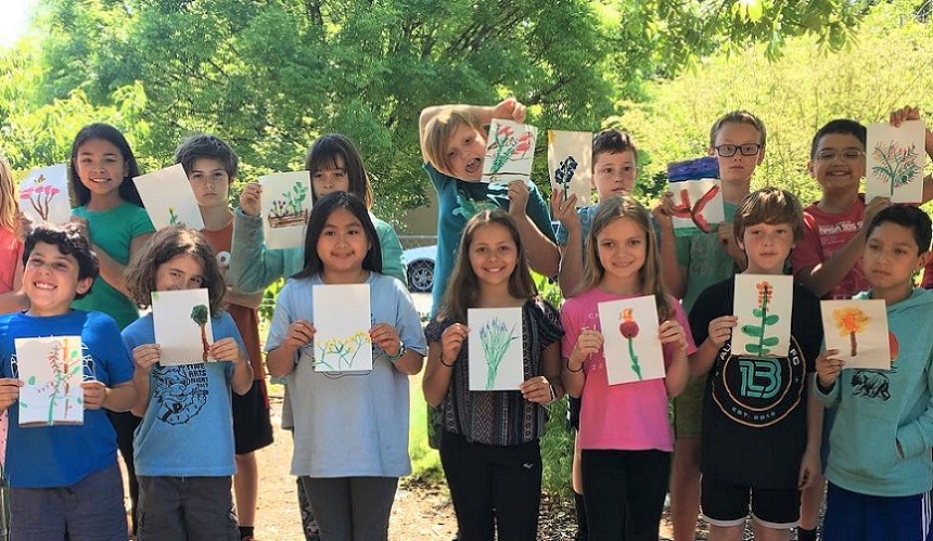 Elementary school students stand outside holding paintings they've made of plants.
