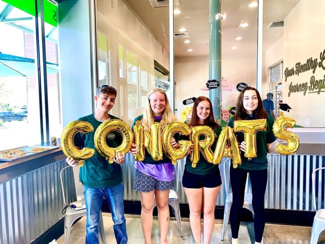 Four graduates in a Sprouts store holding balloons that spell "CONGRATS"