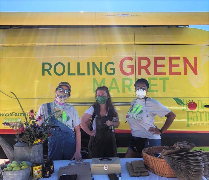 Three women staff a farm stand with a produce van in the background.