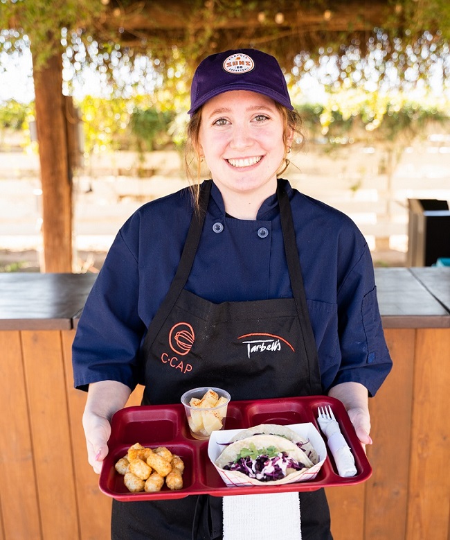 A female chef smiles with a tray full of food.